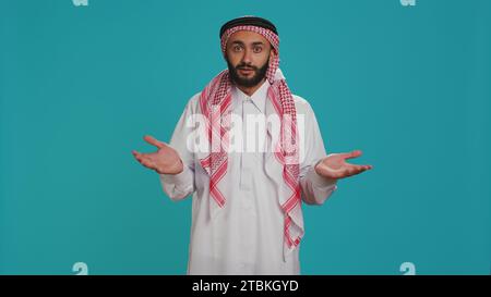 Islamischer Erwachsener, der sich im Studio unsicher und ahnungslos benimmt, das Zeichen ich weiß nicht, und verwirrt über blauem Hintergrund. Junge Person in arabischer Kleidung fühlt sich unsicher und zuckend. Stockfoto
