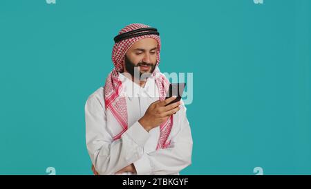 Arabischer Erwachsener in traditioneller Kleidung mit Smartphone, in der Online-Kommunikation in sozialen Medien im Studio. Muslimische Person mit islamischem Kopftuch und Kleidung, im Webbrowser navigieren. Stockfoto