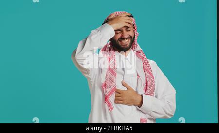 Fröhliche Person lacht über einen Witz im Studio und hat Spaß, während er traditionelle muslimische Kleidung und Kufiyah trägt. Junger arabischer Mann, der mit den Menschen lacht und Glück und Freude ausdrückt. Stockfoto