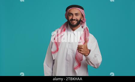 Ein Mann aus dem Nahen Osten zeigt seine Daumen vor der Kamera hoch und zeigt Zustimmung und Positivität, während er traditionelle islamische Kleidung trägt. Junger lächelnder Kerl in nationalem arabischem Kleid, wie Symbol. Stockfoto