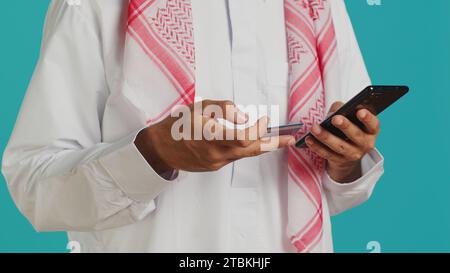 Muslimischer Mann, der Karteninformationen auf der Webseite hinzufügt und Online-Shopping-Sitzungen macht, während er traditionelle islamische Kleidung mit Kopftuch trägt. Person, die Objekte im Webshop oder Bankservice bezahlt Stockfoto