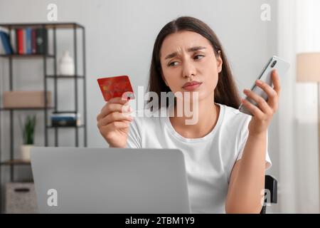 Verwirrte Frau mit Kreditkarte, Smartphone und Laptop zu Hause. Seien Sie vorsichtig – Betrug Stockfoto