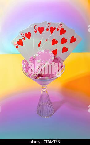 Eine Red Hearts Straight Flush Poker Hand und Pink Poker Chips in einem Martini Trinkglas auf einem hellen Farbverlauf Pastell Hintergrund Stockfoto