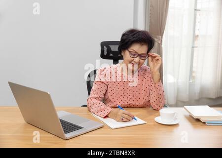 Schreiben Nach Dem Ruhestand. Asiatische Autorin schreibt Details auf dem Buch, während sie zu Hause an einem Laptop arbeitet. Stockfoto