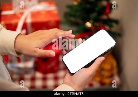 Ein Smartphone-Modell mit weißem Bildschirm befindet sich in der Hand einer Frau über einem unscharfen Hintergrund eines Weihnachtsbaums und Weihnachtsgeschenken in Innenräumen. Menschen und Technologie Stockfoto