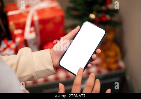 Ein Smartphone-Modell mit weißem Bildschirm befindet sich in der Hand einer Frau über einem unscharfen Hintergrund eines Weihnachtsbaums und Weihnachtsgeschenken in Innenräumen. Menschen und Technologie Stockfoto