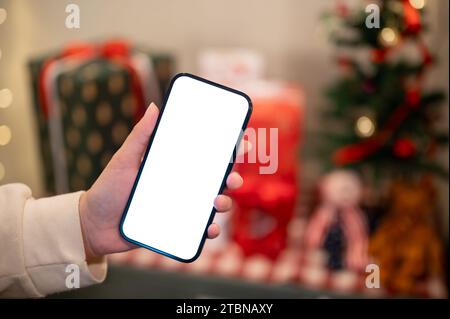 Ein Smartphone-Modell mit weißem Bildschirm befindet sich in der Hand einer Frau über einem unscharfen Hintergrund eines Weihnachtsbaums und Weihnachtsgeschenken in Innenräumen. Menschen und Technologie Stockfoto