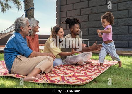 Eine multirassische Familie genießt einen Picknicktag, während ein Kleinkind freudig auf ihre Eltern und Großeltern zuläuft und einen Moment der Familienbindung und Hap festnimmt Stockfoto