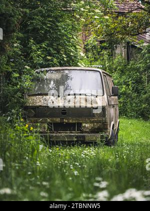 Altes verlassenes Wrack eines Lastwagens in einem Wald, grünes Gras und Bäume, üppiges Laub, Häuser im Hintergrund Stockfoto