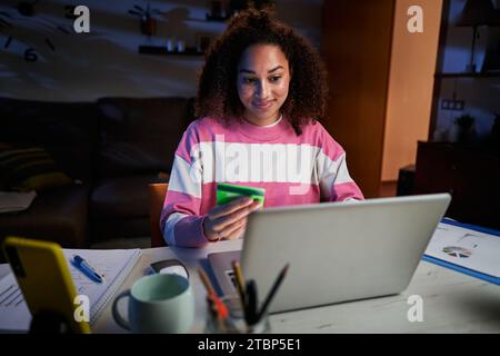 Lächelnde Latina-Frau, die nachts von zu Hause aus Laptop-Shopping im Online-Shop nutzt. Junge Afro-Frau. Stockfoto