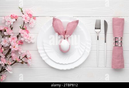 Osterfeiertagstisch mit Ei auf Teller und Besteck, Sakura Blüte, rosa Frühlingsblumen. Weißer Holzhintergrund. Draufsicht, flach. Mittagessen oder Stockfoto