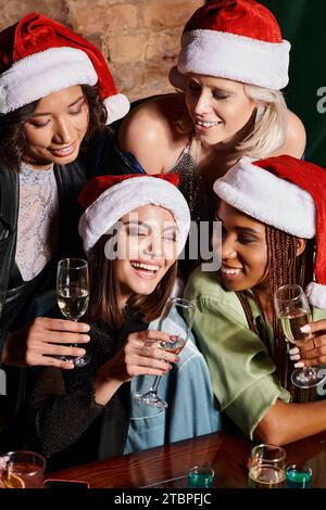 Fröhliche, stilvolle, multiethnische Frauen in Weihnachtshüten mit Sektgläsern in der Bar, festliche Party Stockfoto