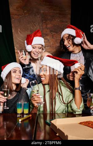 Junge und sorgenfreie multiethnische Freundinnen in Weihnachtshüten mit Champagner, die Spaß in der Bar oder Party haben Stockfoto