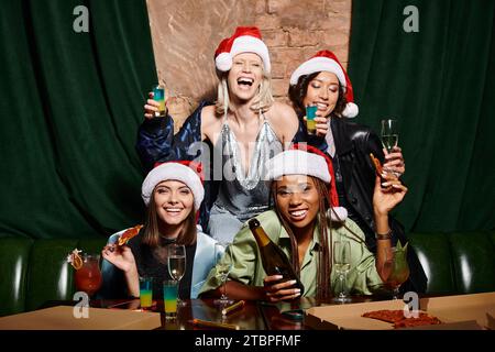 Multirassische Frauen in Weihnachtshüten mit Champagner und Blick auf die Kamera in der Bar, festliche Party Stockfoto