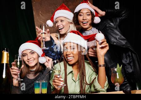 Gruppenporträt von aufgeregten multiethnischen Freundinnen in Weihnachtsmütze mit Sektgläsern in der Bar Stockfoto