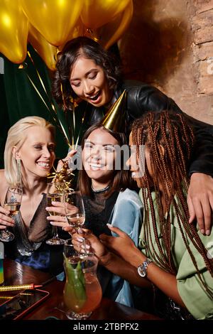 Fröhliche Frau im Partyhut mit Champagner neben multiethnischen Freundinnen und Ballons in der Bar Stockfoto