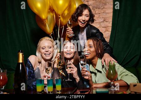 Fröhliche, stilvolle, multiethnische Freundinnen in der Nähe von Cocktails und goldenen festlichen Ballons in der Bar, Geburtstag Stockfoto