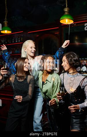 Fröhliche, multiethnische Freundinnen mit Cocktails und festlichen Sparklern, die Spaß in der Bar haben Stockfoto