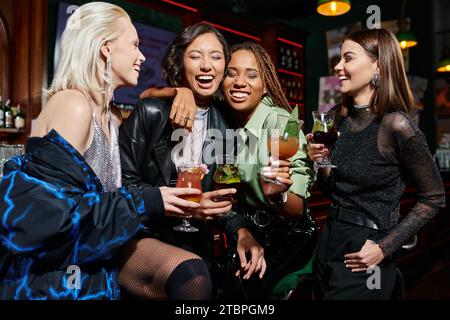 Fröhliche, multiethnische Freundinnen mit Cocktailgläsern, die sich in der Bar umarmen und lachen, Aufregung Stockfoto