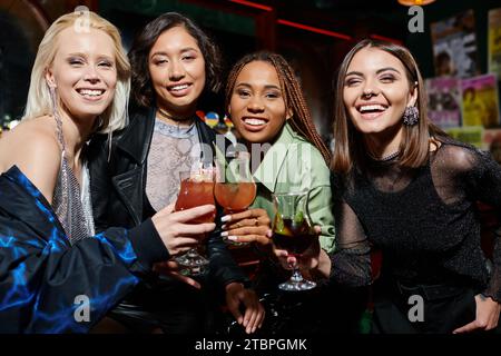 Stilvolle und fröhliche multiethnische Frauen, die Cocktailgläser anklickten und in der Bar vor der Kamera lächeln Stockfoto