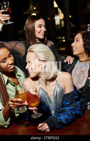 Glückliche, verlockende Frauen, die bei einer Party in der Bar neben multiethnischen Freundinnen Cocktailgläser anklickten Stockfoto