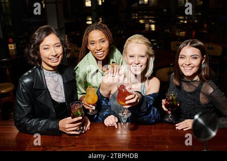 Charmante und fröhliche multiethnische Freundinnen mit Cocktailgläsern, die in der Bar in die Kamera schauen Stockfoto