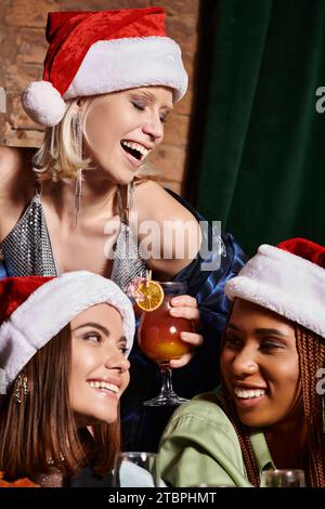 Aufgeregte Frau in Weihnachtsmütze posiert mit leckerem Cocktail in der Nähe von multiethnischen Freundinnen in der Bar Stockfoto