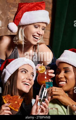 Unbeschwerte multiethnische Freundinnen in Weihnachtshüten, die während der festlichen Party in der Bar die Brille anschlagen Stockfoto