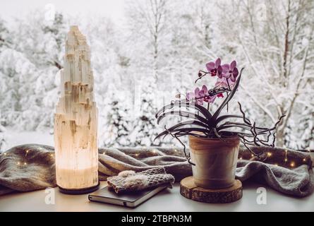 Gemütliches Winterhaus mit elektrischer Turmlampe aus natürlichem Selenit-Kristall beleuchtet auf Fensterbank, grauem Schal und Handschuhen, Papier Notizbuch Orchidee. Stockfoto