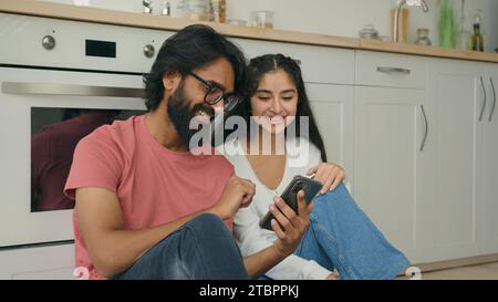 Arabian Indian Interracial Paar Frau Mann sitzt auf warmen Boden in der Küche und sieht lustige Videoinhalte in sozialen Medien, die Online-Komfortlieferung bestellen Stockfoto