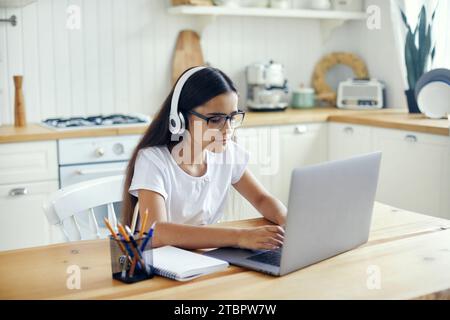 Mädchen im Alter von 12 Jahren in Kopfhörern und Brillen sitzen am Tisch e-Learning, hören Online-Kurse, Audiolektion, erwerben neues Wissen, Fähigkeiten im Internet und Stockfoto