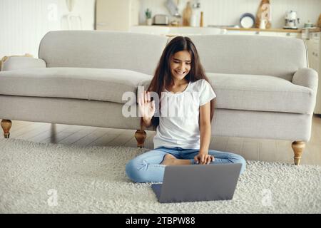 Attraktive nahöstliche Ethnizität vor Teenager 12er Mädchen sitzen auf dem Boden im Wohnzimmer Welle Hand Begrüßung Freund Start Video Veranstaltung Verwenden Sie modernen Laptop. V Stockfoto