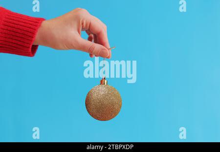 Die Hand einer Frau in einem roten Neujahrspullover hält einen Weihnachtsball an einer Garnschlaufe, blauer Hintergrund. Weihnachtsdekor, goldener Weihnachtsbaumspielzeug Stockfoto