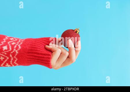 Die Hand einer Frau in einem roten Neujahrspullover hält einen Weihnachtsball, blauer Hintergrund. Weihnachtsdekor, rotes Weihnachtsbaumspielzeug aus nächster Nähe. Banner, Copy Spa Stockfoto