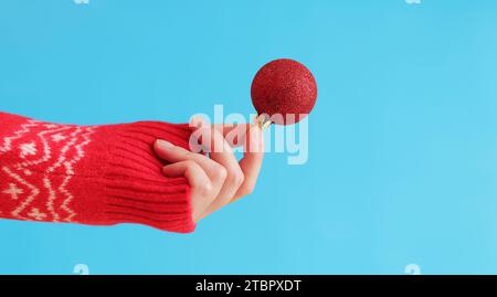 Die Hand einer Frau in einem roten Neujahrspullover hält einen Weihnachtsball, blauer Hintergrund. Weihnachtsdekor, rotes Weihnachtsbaumspielzeug aus nächster Nähe. Banner, Copy Spa Stockfoto