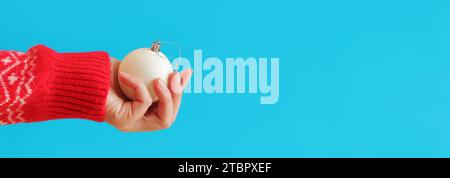 Die Hand einer Frau in einem roten Neujahrspullover hält einen Weihnachtsball, blauer Hintergrund. Weihnachtliche Einrichtung, weißes Weihnachtsbaumspielzeug aus nächster Nähe. Banner, Kopie s Stockfoto