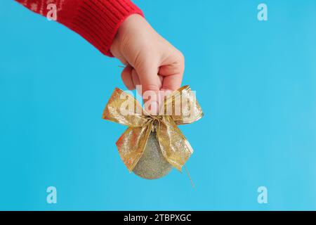 Die Hand einer Frau in einem roten Silvesterpullover hält einen Weihnachtsball mit einer Schleife, blauem Hintergrund. Weihnachtsdekor, Weihnachtsbaumspielzeug in Silber-Gold-Nähe Stockfoto