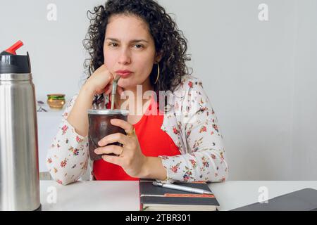 Junge lateinische arbeitende Frau in Lockenwicklern, im Büro sitzend mit Laptop, Telefon und Notebook auf dem Tisch, sie trinkt während ihrer Pause Mate, Bild wi Stockfoto