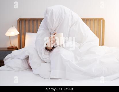 Eine Frau, die morgens aufwacht, eine Decke auflegt und eine Kaffeetasse hält Stockfoto