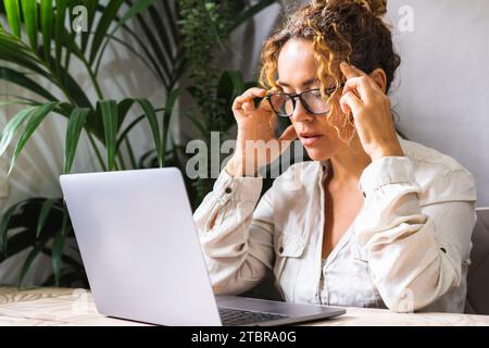 Müde Frau massiert Schläfen, leidet nach der Computerarbeit an Kopfschmerzen, sitzt am Schreibtisch mit Laptop zu Hause, erschöpfte junge Frau mit geschlossenen Augen, die den Kopf berührt, lindert Schmerzen, Migräne Stockfoto