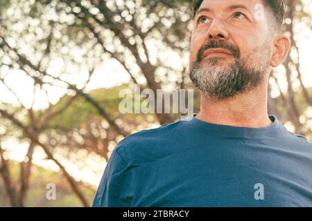 Nahaufnahme Porträt eines selbstbewussten männlichen Erwachsenen, der nach oben blickt. Bärtiger Mann mit blauem T-Shirt und Bäumen im Hintergrund. Menschen und Outdoor Freizeitaktivitäten Konzept Lifestyle. Heiterer Ausdruck im Gesicht. Kopierraum Stockfoto