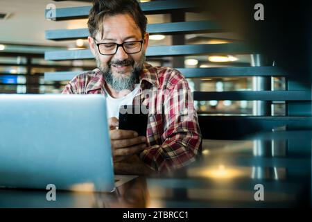 Glücklicher moderner reifer Mann bei der Arbeit in einem kooperierenden Arbeitsplatz, das alternative Büro mit Laptop-Computer und Mobiltelefon zur Kommunikation verwendet. Geräteverbindung. Männliche Erwachsene lächeln und Smartphone-App in Innenräumen verwenden Stockfoto