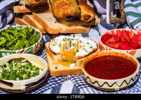 Rustikale Komposition mit Würstchenbrötchen, Spiegelei auf Toast, verschiedenen Schüsseln mit Sauce und gehacktem Gemüse. Stockfoto