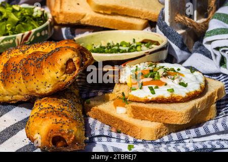 Rustikale Komposition mit Würstchenbrötchen, Spiegelei auf Toast, verschiedenen Schüsseln mit Sauce und gehacktem Gemüse. Stockfoto