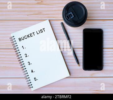 Das Wort „Eimer List“ ist auf einem Notizblock mit Kaffee, Stift und Smartphone-Hintergrund auf einem Holztisch geschrieben. Stockfoto