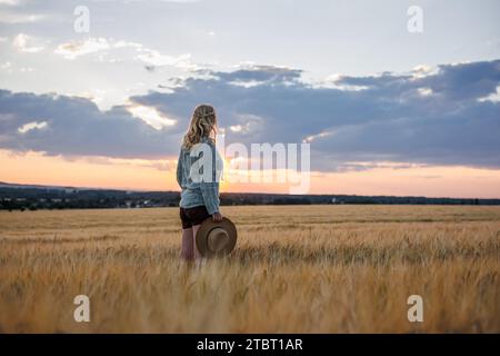 Zufriedene Farmerinnen schauen sich Gerstenfelder auf bewirtschaftetem Land an. Frau mit Strohhut, die während des Sonnenuntergangs auf dem landwirtschaftlichen Feld steht Stockfoto