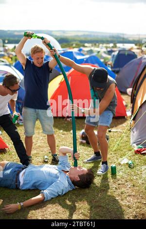 Mann, Freunde und Bierpfeife auf Outdoor Festival, Camp oder Party zusammen mit Alkohol auf grünem Gras oder Feld. Gruppe von Leuten, die in den Schlauch trinken Stockfoto