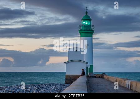 Frankreich, Normandie, Le Tréport, Leuchtturm mit Leuchtfeuer in Grün, pastellfarbene Abendatmosphäre mit Wolken, Paare sitzen auf der Kaimauer, Hafenmauer, Fußweg, Kieselstrand und Meer Stockfoto