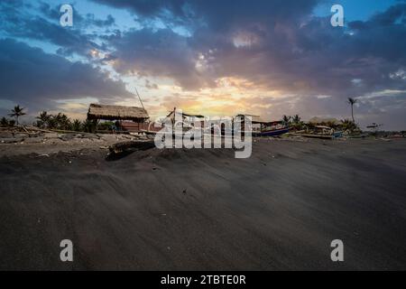 Traditionelles Fischerboot Jukung, auf einem schwarzen Sandstrand am Meer, Landschaft bei Sonnenuntergang am Meer auf der tropischen Insel Sanur, Bali, Indonesien Stockfoto