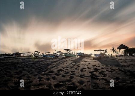 Traditionelles Fischerboot Jukung, an einem schwarzen Sandstrand direkt am Meer, abendliche Landschaft am Meer auf der tropischen Insel Sanur, Bali, Indonesien Stockfoto